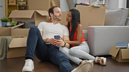 Fototapeta na wymiar Beautiful couple sitting on floor together using smartphone smiling at new home