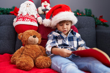 Obraz na płótnie Canvas Adorable hispanic boy reading book sitting on sofa by christmas decoration at home