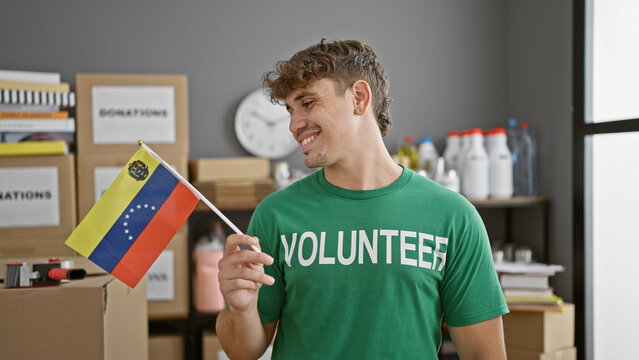 Handsome young hispanic man, a smiling volunteer, brimming with patriotic pride, holds venezuelan flag at vibrant charity center, standing as a pillar of unity and social support.