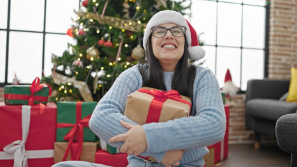 Obraz na płótnie Canvas Young hispanic woman smiling wearing christmas hat holding present at home