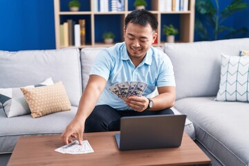 Young chinese man using laptop counting dollars at home