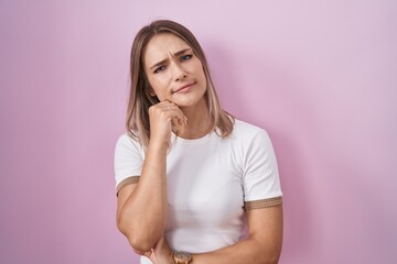 Blonde caucasian woman standing over pink background with hand on chin thinking about question, pensive expression. smiling and thoughtful face. doubt concept.