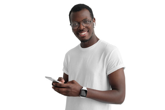 Young african american man looking and smiling at the camera, using mobile phone, listening to music with white wireless earphones