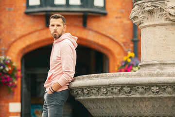 Young bearded romantic man wearing pink trendy hoodie, gray jeans posing over brick house with flowers on backgound. Old classical fontain. Outdoor shot