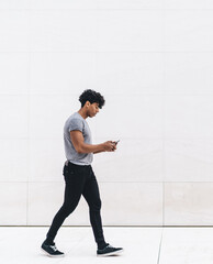 Handsome young curly man in casual outfit browsing smartphone while walking on marble passage