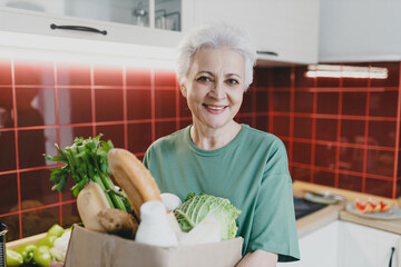 Senior female holding carton box with food products and vegetables after ordering home delivery from grocery store posing in kitchen, going ready to cook delicious healthy family dinner
