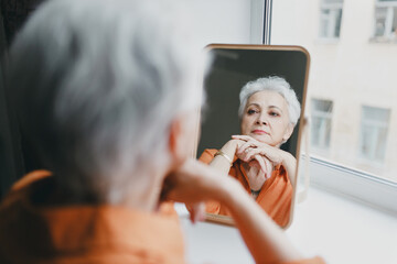 Senior elderly female with gray hair and wrinkles looking in mirror, upset with aging process of...