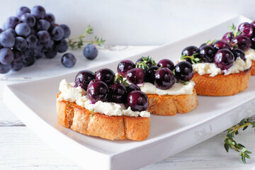 Crostini appetizers with roasted grapes and whipped feta cheese. Close up table scene against a white background. Party food concept.