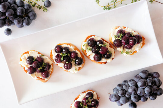Crostini appetizers with roasted grapes and whipped feta cheese. Overhead view table scene against a white marble background. Party food concept.