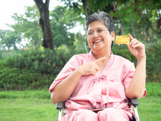 Happy Asian senior woman in pink patient uniform holding credit card and sitting on wheelchair at park. Elderly health care.