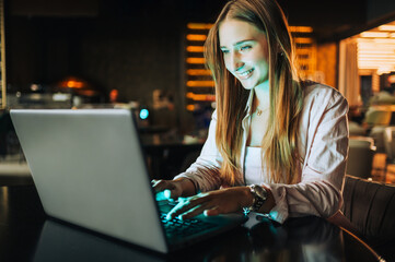 A cheerful young female entrepreneur is sitting in a cafe and typing on a laptop in the dark while smiling at the laptop.