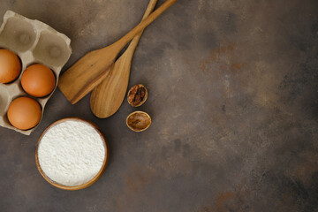 Kitchen utensils and ingredients for baking, spatula, eggs, flour on brown background.