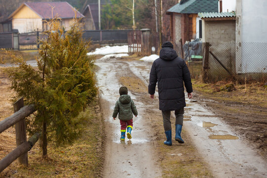 Father and son walking in the fresh air in rubber boots on the puddles and mud after the rain. Little child holding hand of a man