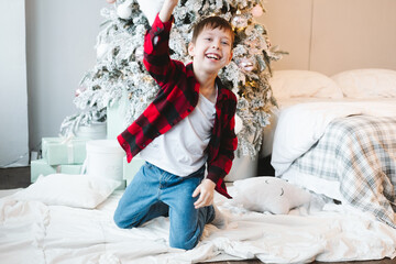 A boy in a red checkered shirt sits by the Christmas tree with gifts on the floor and fights with a pillow