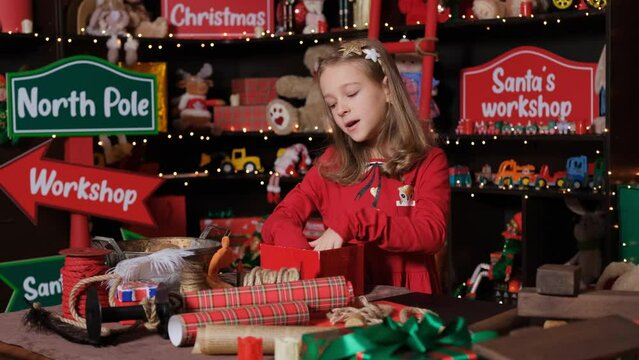 Little Girl Santa helper Elf packing Christmas gift at workshop, slow motion. Lots of toys and gift wrapping materials around her