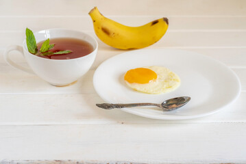Cup of mint tea, plate with sunny side up egg on white wooden background. Breakfast food.