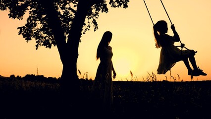 Mother helping little girl to swing dark silhouettes in rural park at sunset
