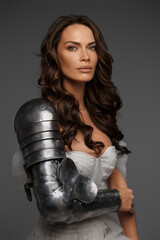 Beautiful middle-aged model in white dress and knight's armor poses gracefully on gray