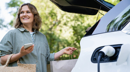 Woman with shopping bag next to a charging electric car in the yard of a country house