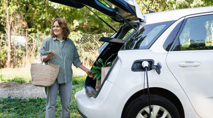 Woman with shopping bag next to a charging electric car in the yard of a country house