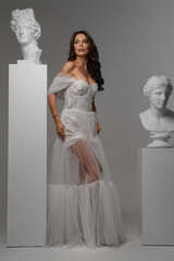 Elegant woman in white gown juxtaposed with classical Greek statues in a studio setting