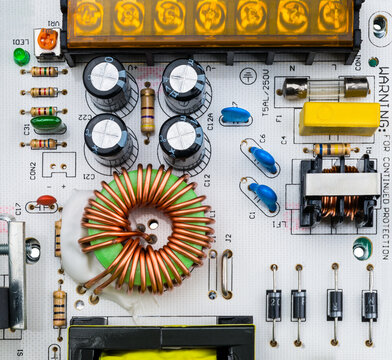 Close-up of electronic components on white printed circuit board. Toroidal coil and various capacitors, electrical fuse and resistor trimmer or diodes on power supply inverter PCB with screw terminal.