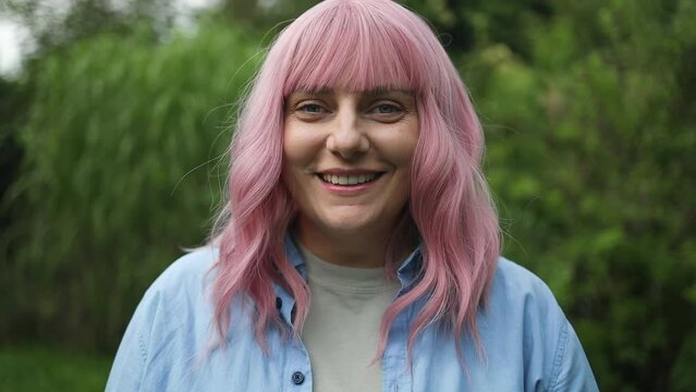 Close-up portrait of attractive pink-haired woman in bright colorful casual clothes smiling at the camera outside in a backyard or park.