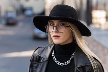 Portrait of an attractive and young blonde woman in a black hat and a leather trench coat while walking around the city. The concept of style and fashion