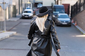 An attractive and young woman in a black hat and a leather trench coat walks around the city on an autumn day. View from the back