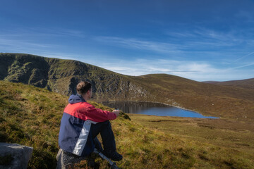 Naklejka premium Men sitting on a rock and drinking coffee or tea from thermos. Scenic view on Lough Ouler and Tonelagee Mountain, hiking in Wicklow Mountains, Ireland