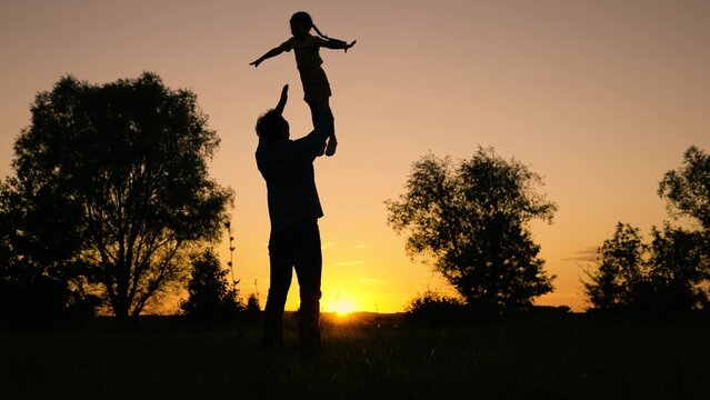 Dad plays with his daughter, throws up child with his hands in sky, outdoors. Family game concept. Child, superhero, fly. Happy Father, child, little girl have fun together in park against sun. Kid