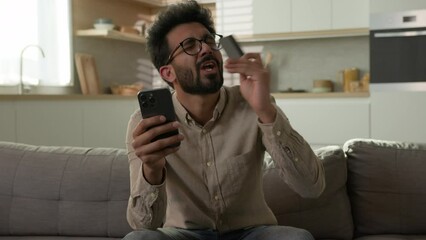 Happy Indian smiling man shopper buyer client paying online with mobile phone in home kitchen internet web store pay in e-banking approved money transfer joyful Arabian ethnic guy kissing credit card