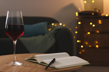Glass of red wine and open notebook with pen on wooden table in room. Relax at home