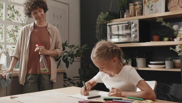 Slowmo of proud mother watching her creative daughter drawing picture while cleaning kitchen