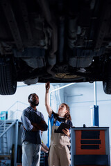 Engineer in car service uses tablet to calculate invoice after fixing broken automobile tires. Worker uses device to inform client of final costs after mending vehicle suspended on overhead lift