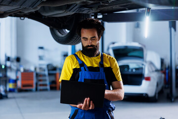 Mechanic in car service uses laptop to write invoice after repairing client automobile wheels. Productive garage professional calculating final costs after refurbishing customer vehicle