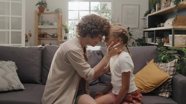 Medium slowmo of loving mother sitting on couch with her preteen daughter forehead to forehead, stroking her face