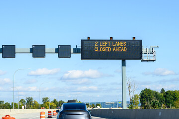 Overhead electronic highway sign, 2 left lanes closed ahead, on a sunny blue sky day
