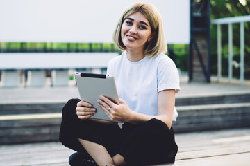 Pleased adult female surfing tablet sitting on stage in street