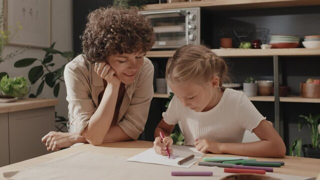Caucasian woman standing at table in kitchen watching her preteen daughter drawing picture