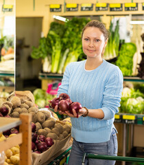 Positive middle-aged woman purchaser taking red onions at the counter in grocery store