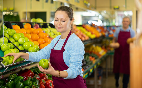 Skillful female merchandiser in uniform checking green apples before selling in grocery supermarket