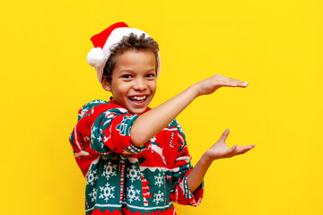 funny african american boy in christmas clothes holds empty hands in his hands on yellow isolated background