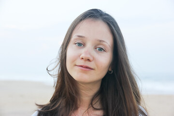 Portrait of a beautiful young woman with clear soft skin. Relaxed girl posing on camera. Dreamy lady smiling looking at a camera. Brown haired female tourist with light eyes on the ocean sea coast.