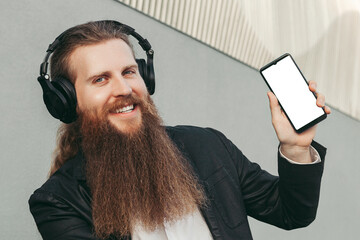 Man european young producer cheerful positive with long beard in jacket in headphones with phone in hands, looking at camera, listening to music and showing phone screen, grey background ,copyspace