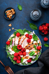 Tasty salad with raspberries, grilled chicken, cheese, onion, walnuts, spinach and arugula, blue table background, top view