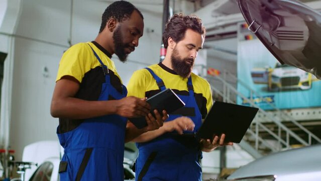 Mechanics in car service ordering new parts for damaged vehicle using laptop and tablet. Coworkers looking online for components to replace old ones in malfunctioning automobile
