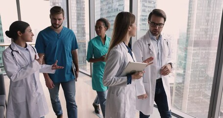 Five motivated multinational therapists, professional medical workers in uniforms walk along clinic hallway and talking, going to group meeting conference or concilium. Profession, medicine, council
