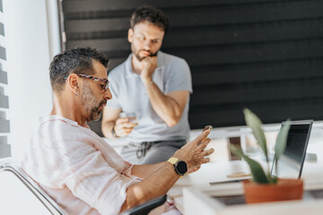 Fototapeta premium Different age coworkers working together at the office. Senior businessman reading mail from client on smart phone to his colleague.