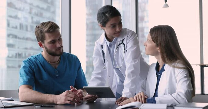 Female general practitioner talks to colleagues, provide help, assist with new medical app software use tablet, discuss patient disease treatment plan gathered in office. Cooperation, tech, medicare